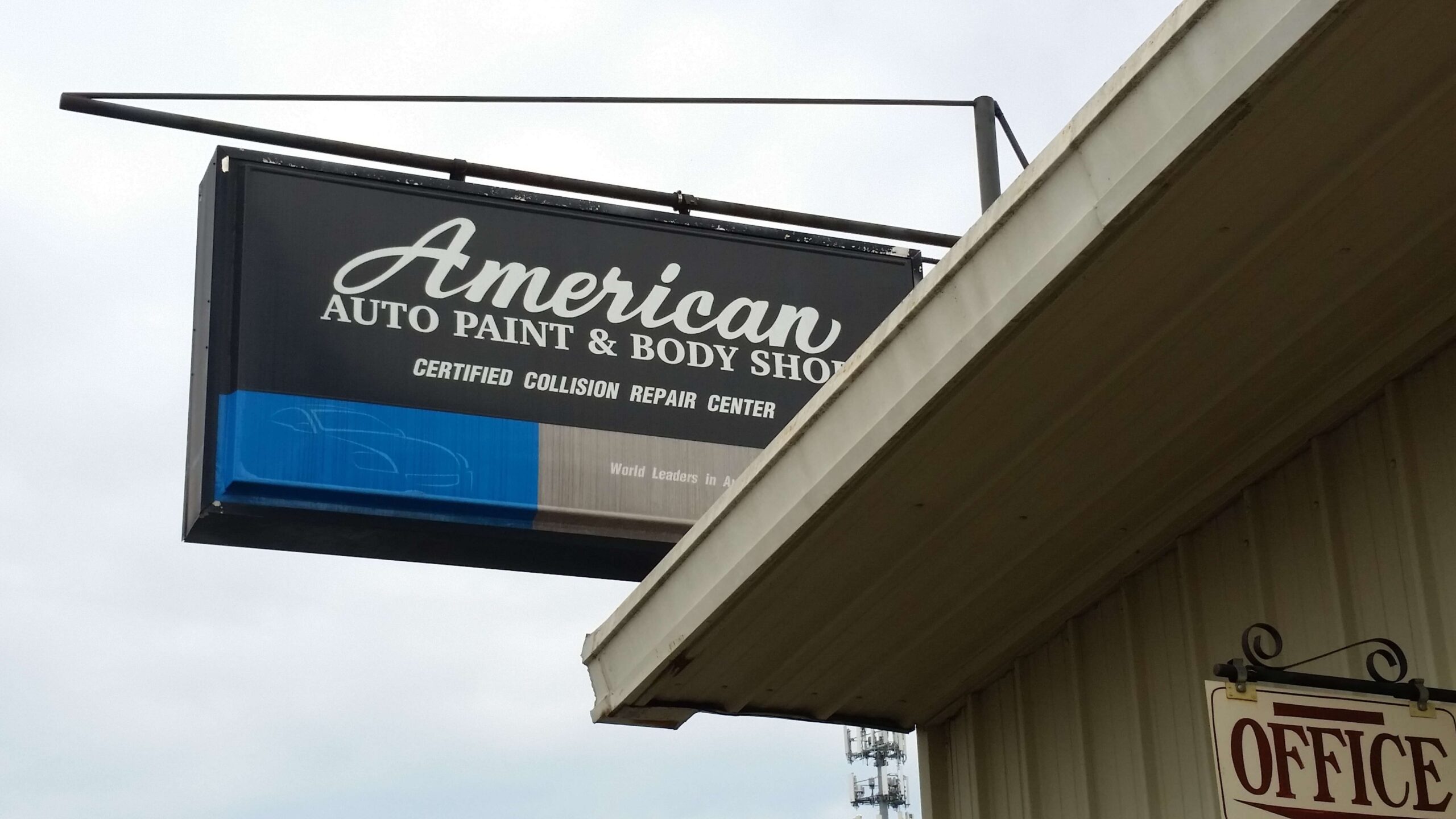 Sign for "American Auto Paint & Body Shop, Certified Collision Repair Center" on a building corner, with an "Office" sign below.