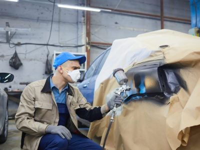 A person in a beige jacket and blue cap spray paints a car door in a workshop. The door is covered with masking paper.