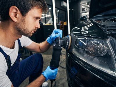 A mechanic in blue gloves polishes a car's headlight using a power buffer in a garage.