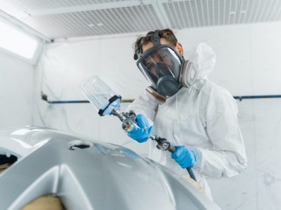 Person in protective gear spray paints a car part in a workshop.