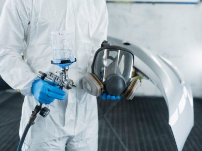 Person in a protective suit holds a spray gun and a respirator mask in a workshop with a vehicle part in the background.