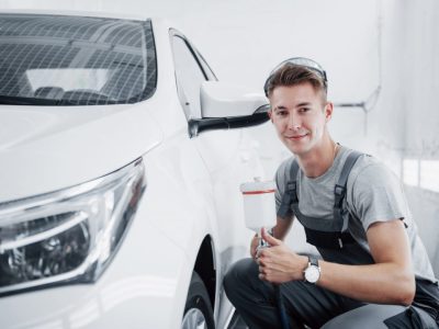 A person in overalls and safety glasses kneels beside a white car in a garage, holding a paint sprayer.